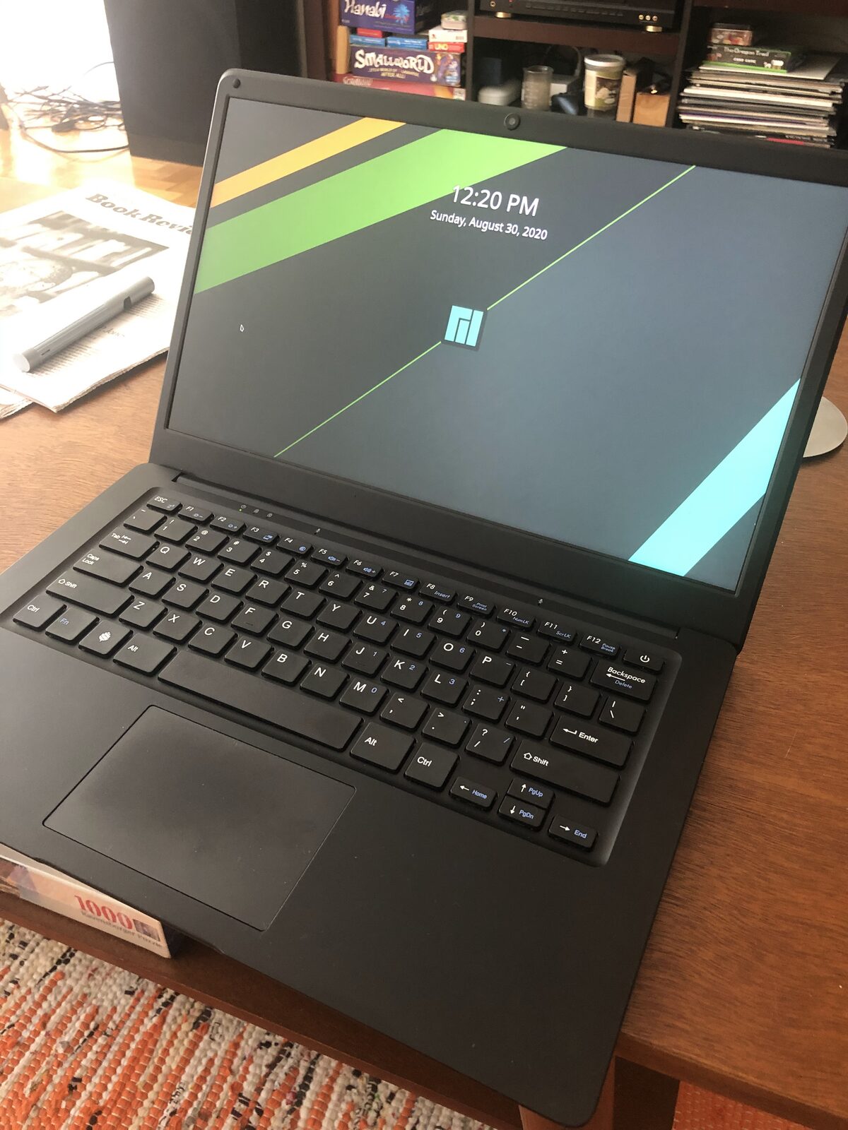 The Pinebook Pro open on a coffee table, showing its lock screen, with bookshelves and a newspaper alongside